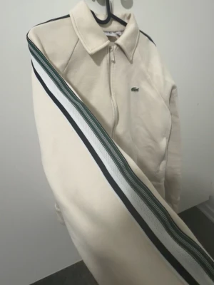 Lacoste - Snygg Lacoste beige jacka med dragkedja och krage. Jackan har en grön och vit rand längs ärmen och en liten logga på bröstet. Perfekt för dig som gillar sportig och clean stil. Storleken är L men den passar som M eftersom koftan kommer en storlek mindre. Priset kan diskuteras