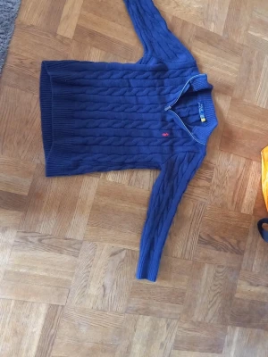 Mörkblå kabelstickad tröja Polo Ralph Lauren - Mörkblå kabelstickad tröja från Polo Ralph Lauren med röd broderad logga på bröstet. Tröjan har rund halsringning och ribbade muddar vid ärmslut och nederkant. Klassisk och tidlös design i mjukt material.