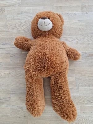 Stor brun mjuk nallebjörn - Säljer en stor och fluffig nallebjörn i mjuk plysch. Björnen är ljusbrun med ett stort runt huvud, svart broderade ögon och en vit nos med brunt broderad mun och näsa. Perfekt som mysig inredningsdetalj eller present till någon du gillar.