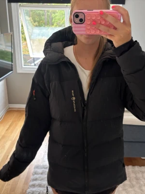 Svart dunjacka från sail racing  - Säljer en svart dunjacka från sail racing med huva och dragkedja framtill. Jackan har en sportig look, diskret logga på bröstet och en ficka med dragkedja på ärmen. Perfekt för kalla vinterdagar och riktigt skön att bära.