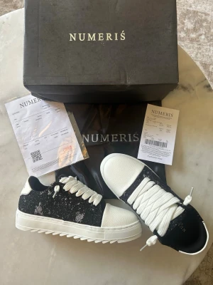 Svarta och vita sneakers från Numeris - Snygga sneakers från Numeris i svart och vitt med chunky sula och breda vita skosnören. Ovandelen har en cool svart bas med vita stänkdetaljer och vit tå samt hälparti i läderlook. Perfekta för dig som vill sticka ut med din stil.
