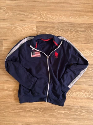 Marinblå ziptröja från Ralph Lauren - Marinblå ziptröja med ståkrage och vita detaljer längs ärmarna. Broderad USA-flagga och texten 'USA R. Lauren' på bröstet samt röd Polo-logga. Perfekt för en sportig och avslappnad stil.
