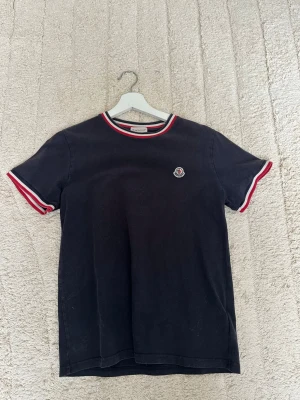 Svart Moncler t-shirt med rödvit kant - Snygg svart t-shirt från Moncler med korta ärmar och klassisk rund hals. T-shirten har röda och vita detaljer på ärmslut och halsringning samt Moncler-logga på bröstet. Tillverkad i mjuk bomull . Storlek 14A passar S . Passar mig inte längre . Ny pris 1500