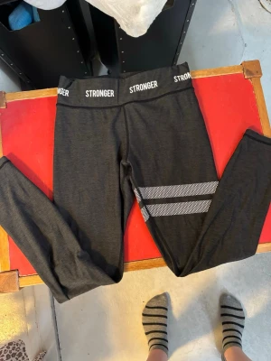 Svarta tights från Stronger - Snygga svarta träningstights från Stronger med vita logotyper i midjan och två vita randiga detaljer på ena benet. Tightsen har hög midja och är tillverkade i ett stretchigt material som passar perfekt för gymmet eller löprundan. St S, används fåtal gånger och i väldigt bra skick!