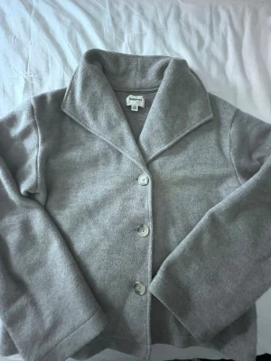 Ljusgrå kappa från Kappahl - Stilren ljusgrå kappa från Kappahl med klassisk krage och tre knappar framtill. Kappan har raka ärmar och är tillverkad i ett mjukt tyg som ger en clean look. Perfekt för dig som gillar enkel och tidlös stil.