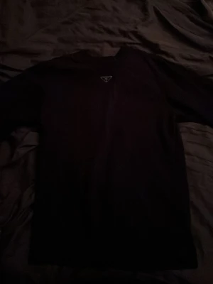 Svart t-shirt från Prada - Snygg svart t-shirt från Prada med den klassiska metalltriangeln på bröstet. Enkel och stilren design som passar till allt. Tillverkad i mjukt material för skön känsla och har normal passform med korta ärmar.