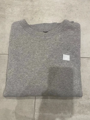 Grå hoodie med broderad patch - Snygg grå hoodie med en broderad patch framtill som har ett minimalistiskt ansikte. Hoodien har klassisk passform. Unisex.