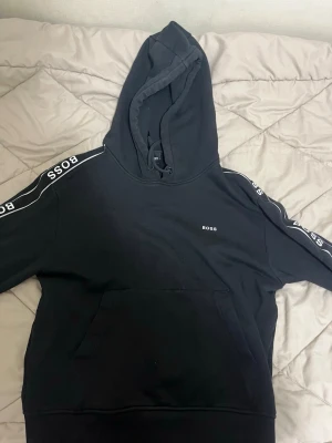 Svart hoodie från BOSS  - Säljer en svart hoodie från BOSS med vit logga på bröstet och längs båda ärmarna. Klassisk känguruficka framtill, huva med dragsko och ribbade muddar. Snygg streetstil och skön passform, perfekt till jeans eller joggers.