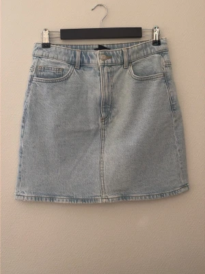 Ljusblå jeanskjol med fickor - Säljer en klassisk ljusblå jeanskjol i kort modell med fem fickor och knapp- samt dragkedjestängning framtill. Kjolen har en rak passform och är tillverkad i bomull, perfekt för en avslappnad och trendig look.