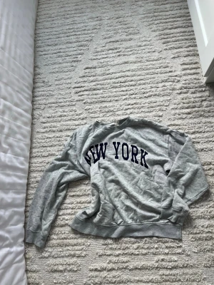 Grå sweatshirt med New York-tryck - Säljer en grå sweatshirt från H&M med stort mörkblått 'NEW YORK'-tryck på bröstet. Tröjan har rund halsringning, långa ärmar och ribbade muddar vid ärmslut och nederkant. Perfekt för en chill och avslappnad stil.