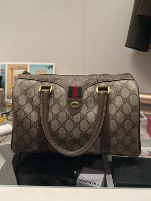 Gucci brun monogram handväska - Snygg handväska från Gucci i klassiskt beige-brunt monogramtyg med mörkbruna läderdetaljer. Väskan har dubbla handtag, guldfärgade metalldetaljer och den ikoniska gröna och röda randen med GG-logga framtill. Perfekt för dig som gillar lyxig och tidlös design.