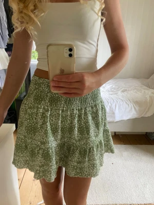 Grön blommig kjol från Gina Tricot - Supersöt grön kortkjol från Gina Tricot med vitt blommönster och volangdetalj. Kjolen har smock i midjan för en bekväm passform och är perfekt för varma dagar. Lätt och luftig känsla.