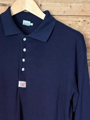 Vintage Levi’s 90s waffle thermal polo navy XL - Vintage Levi’s långärmad polo 90-tal XL  Äldre Levi’s-polo i mörk marinblå 100% bomull med kraftig waffle/thermal-struktur. Klassisk krage, fyra knappar, ribbade muddar och liten “Tried and True”-patch framtill. Avslappnad 90s-passform och en ovanligare Levi’s-modell.  Fint vintagebruksskick.  XL | Bröst 53 cm | Axlar 53 cm | Ärm 58 cm | Längd 74 cm