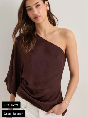 Brun oneshoulder topp i satin - Snygg brun oneshoulder topp i glansigt satinmaterial. Toppen har en asymmetrisk design med ena axeln bar och ett ledigt fall. Perfekt för dig som vill sticka ut med en trendig och elegant look.