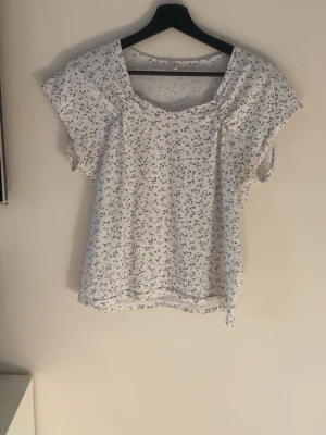Vit blommig blus med kort ärm - Söt vit blus med småblommigt mönster i blått och lila. Blusen har korta ärmar och en rundad halsringning. Perfekt för vår och sommar, lätt och luftig i materialet.