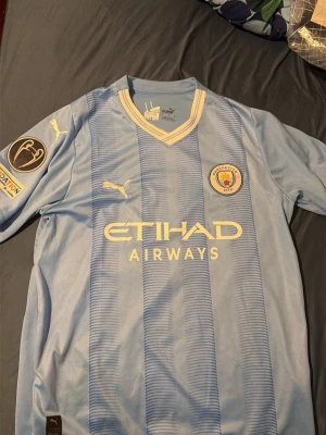 Manchester City matchtröja Puma - Säljer en ljusblå Manchester City matchtröja från Puma med vita detaljer och klubbmärke på bröstet. Tröjan har V-ringning med guldkant, Etihad Airways-tryck på framsidan och Champions League-märke på ärmen. Perfekt för fotbollsfans!