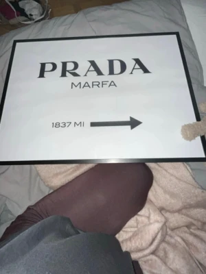 Prada Marfa tavla i svartvitt - Snygg svartvit tavla med texten 'PRADA MARFA' och en pil med avståndet 1837 MI. Tavlan har en svart ram och modern design, perfekt som inredningsdetalj för dig som gillar mode och stilrena posters. Både affischen och ram ingår
