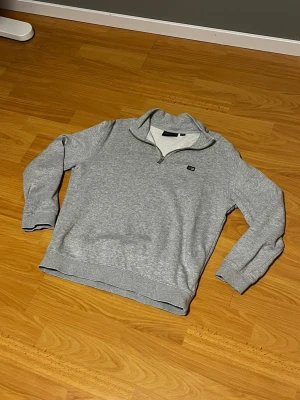 Grå halvzip hoodie från Lexington - Snygg grå halvzip hoodie från Lexington med liten logga på bröstet. Tillverkad i mjukt bomullsmaterial med borstad insida för extra komfort. Klassisk passform, ribbade muddar och krage med dragkedja. Perfekt för en avslappnad och stilren look.