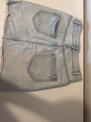Ljusblå jeanskjol med slitningar - Säljer en ljusblå jeanskjol med rå kant och slitna detaljer framtill och baktill. Kjolen har klassiska fickor både fram och bak samt bälteshällor i midjan. Perfekt för dig som gillar en avslappnad och trendig stil.
