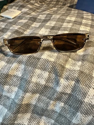 Gucci solglasögon med guldiga detaljer - Snygga solglasögon från Gucci med rektangulära, mörkbruna glas och tunna guldiga metallbågar. Glasögonen har en stilren och modern design med diskreta logodetaljer på skalmarna. Perfekta för dig som vill ha en exklusiv accessoar till sommaren.