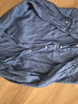 Blå linneskjorta från Riley - Säljer en stilren blå skjorta från Riley i mjukt linne. Skjortan har klassisk krage, vita knappar framtill och lång ärm. Perfekt för dig som gillar en avslappnad men ändå snygg look.