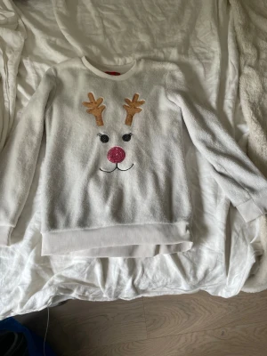 Vit jultröja  - Mysig vit tröja med ett gulligt renansikte framtill, glittrig röd nos och bruna horn. Tröjan har långa ärmar, ribbade muddar och är perfekt för julens alla tillfällen. Materialet ser ut att vara mjukt och fleece-liknande.