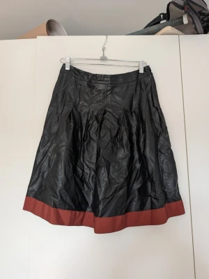 Svart långkjol med brun kant - Säljer en svart långkjol från Klub med bred brun kant längst ner. Kjolen har en lätt glansig yta och är gjord i ett material som liknar skinn. Perfekt för dig som vill ha en edgy och unik look.