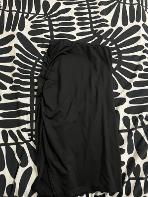 Svart omlottkjol med slits till barn  - Säljer en svart omlottkjol i mjukt tyg med snygg slits på sidan. Kjolen har en draperad detalj längs ena höften som ger en cool och trendig look. Perfekt för dig som gillar stilrena plagg med en twist.