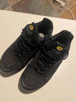 Nike Air Max Plus svarta sneakers - Säljer ett par svarta Nike Air Max Plus sneakers med TN-logga på plösen. Skorna har en sportig design med svarta detaljer och reflekterande inslag. Ovandelen är i mesh och syntetmaterial, och sulan är robust med synlig Air-dämpning.