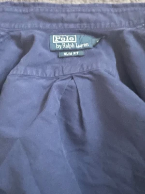 Mörkblå skjorta från Polo Ralph Lauren - Säljer en mörkblå långärmad skjorta från Polo Ralph Lauren i slim fit-modell. Skjortan har klassisk krage, vita knappar framtill och den ikoniska röda logotypen broderad på bröstet. Perfekt för en stilren och avslappnad look.