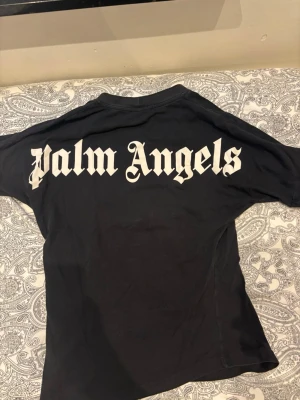 Svart Palm Angels t-shirt - Svart t-shirt från Palm Angels med stor vit logga i gotisk stil på ryggen och mindre logga vid halsen fram. Klassisk passform och rund halsringning. Perfekt för dig som gillar streetwear och vill sticka ut med en ikonisk design.