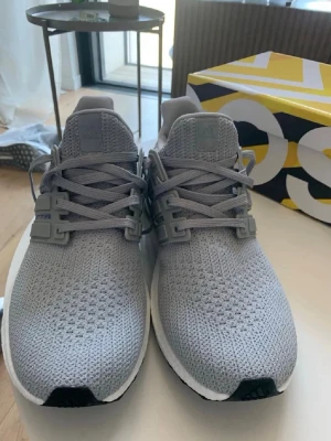 Grå Adidas Ultraboost sneakers - Snygga grå Adidas Ultraboost sneakers med stickad ovandel i mesh för maximal komfort och andningsförmåga. Skorna har klassiska tre ränder på sidan, vit mellansula och svart yttersula. Perfekta för dig som gillar stilrena och sportiga sneakers.
