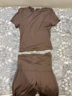 Brunt loungewear-set i mjukt tyg - Mysigt loungewear-set i mjuk, stretchig brun trikå. Setet består av en kortärmad t-shirt med rund hals och ett par vida byxor med bred, hög midja. Perfekt för chill hemma eller avslappnade dagar. Materialet känns lent mot huden och ger en avslappnad look.