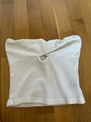 Vit tubtopp med twistad detalj - Säljer en vit tubtopp med twistad detalj framtill och liten öppning under bysten. Toppen är ärmlös och har en lätt strukturerad yta i ett mjukt material. Perfekt för sommaren och enkel att styla till olika looks.