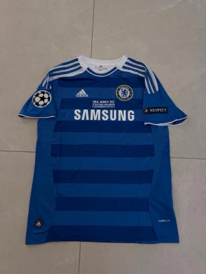 Chelsea FC blå matchtröja Adidas - Chelsea FC matchtröja i blått med mörkare blå ränder, vita detaljer och tryck. Adidas-logga, Champions League-märke och Samsung som sponsor på bröstet. Tröjan har klubbmärke, vita ränder på axlarna och är tillverkad i lätt, ventilerande material.