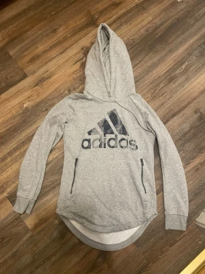 Grå hoodie från Adidas stl S med dragkedjor - Snygg grå hoodie från Adidas med stor logga på bröstet och två praktiska fickor med dragkedja. Hoodien har långa ärmar, ribbade muddar och längre bakstycke för extra stil. Perfekt för chill eller träning.