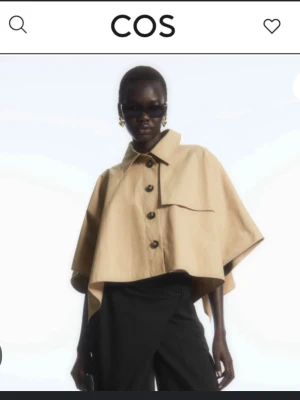 Kort beige trenchcoat från COS - Stilren kort trenchcoat från COS i beige bomullsmix. Jackan har vida, halvlånga ärmar, klassisk krage och stora knappar framtill. Oversized och boxig passform ger en modern look. Perfekt för lager-på-lager och trendiga outfits. Har använts två gånger!