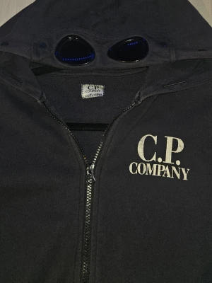 Mörkblå C.P. Company Hoodie - Äkta Mörkblå C.P. Company Hoodie med sin väldigt unik och coola design men glasögon i huvan och med ett snyggt tryck på bröstet. Storlek S. OPS! Jag kan bara skicka med PostNord.
