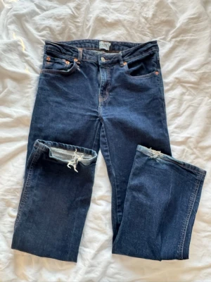 Mörkblå raka jeans med slitning - Säljer ett par mörkblå jeans med rak passform och slitning vid ena knät. Klassisk femficksmodell med knappgylf och detaljer i kopparfärg. Jeansen har bakfickor med lock och knapp. Perfekta för en avslappnad och trendig look.