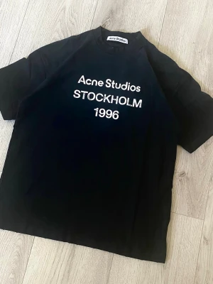 Svart Acne Studios t-shirt - Säljer en svart t-shirt från Acne Studios med vit text på bröstet där det står 'Acne Studios STOCKHOLM 1996'. Klassisk passform och rund halsringning. Perfekt till jeans eller shorts för en clean och stilren look.