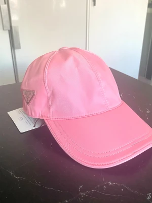 Rosa keps från Prada  - Snygg rosa keps från Prada i glansigt nylonmaterial. Klassisk modell med böjd skärm och justerbart spänne bak. Prada-logga på sidan och präglad text bak. Perfekt accessoar för en trendig look.
