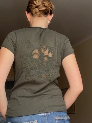 Olivgrön t-shirt med mönstrad rygg - Säljer en olivgrön t-shirt med korta ärmar och v-ringning från Zadig & Voltaire. Baksidan har en unik mönstrad sliten detalj av en döskalle. Perfekt för dig som gillar en avslappnad och cool stil. Tillverkad i mjuk bomull. Snygg passform o logga vid vänstra armen, få tecken på användhet 