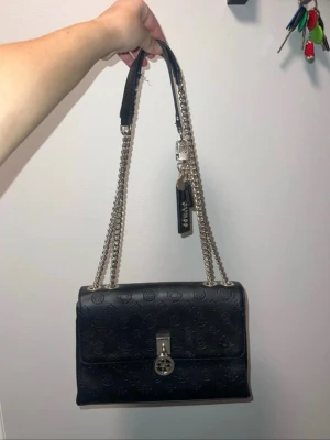 Svart handväska med kedjerem från Guess - Snygg svart handväska från Guess i präglat konstläder med logomönster. Väskan har silverfärgade kedjeremmar och ett dekorativt lås framtill. Guess-tag i metall och läder hänger på sidan. Perfekt storlek för det viktigaste.