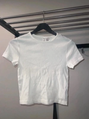 Vit ribbad t-shirt från Divided H&M - Säljer en enkel och fräsch vit ribbad t-shirt från Divided. T-shirten har korta ärmar, rund halsringning och en tight passform. Perfekt att styla med jeans eller kjol för en clean look. Finns en liten fläck på ena axeln.
