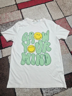 Vit t-shirt med grön text och smileys - Vit t-shirt med stort tryck i grönt och gult på framsidan där det står 'Grow Your Mind' och två smileys. T-shirten har rund hals och avslappnad passform, perfekt för en chill och färgglad stil. storlek 158