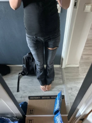 Svarta utsvängda jeans med slitningar - Snygga svarta jeans med utsvängda ben och slitningar på knäna och låret. Jeansen har stora fickor fram och klassiska bakfickor. Perfekta för dig som gillar en edgy och trendig stil. Materialet är denim med en tvättad finish.