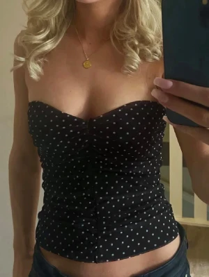Svart prickig bandeau tubtopp - Säljer en svart bandeau tubtopp med vita prickar och rynkade detaljer framtill. Toppen är axelbandslös och har en figurnära passform, perfekt för dig som vill ha en trendig och enkel look. Materialet känns mjukt och stretchigt.