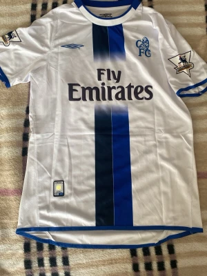 Chelsea Lampard 8 fotbollströja  - Chelsea FC matchtröja i vitt med blå vertikal rand, Lampard 8 på ryggen och Fly Emirates som sponsor. Tillverkad av Umbro med Premier League-märken på ärmarna. Klassisk fotbollströja i lätt och ventilerande material, perfekt för fans och samlare.