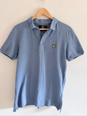 Blå pikétröja från Lyle & Scott - Blå pikétröja från Lyle & Scott med klassisk krage, knappslå och broderad gul logga på bröstet. Tillverkad i mjuk bomull och har korta ärmar för en stilren och avslappnad look.