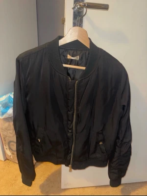 Svart bomberjacka med dragkedja - Snygg svart bomberjacka med klassisk ribbad krage och muddar. Jackan har dragkedja framtill och två fickor med knappstängning. Tillverkad i ett glansigt material som ger en cool look. Perfekt för dig som vill ha en stilren och enkel jacka.